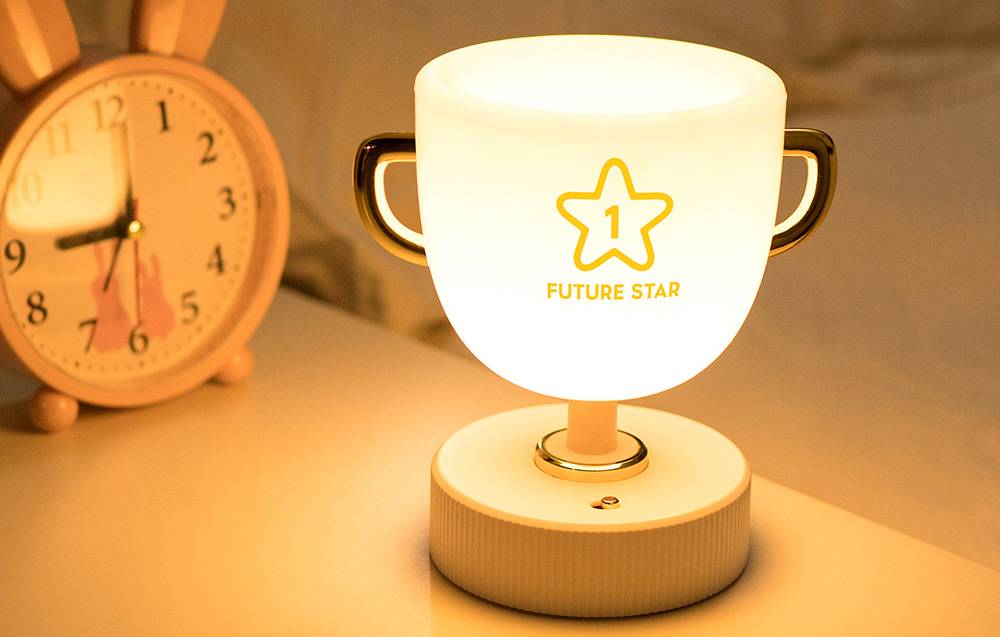 SuperFire W-006 Champion Light children's night light glowing with white light. The device is placed on a bedside table next to a rabbit-shaped alarm clock.